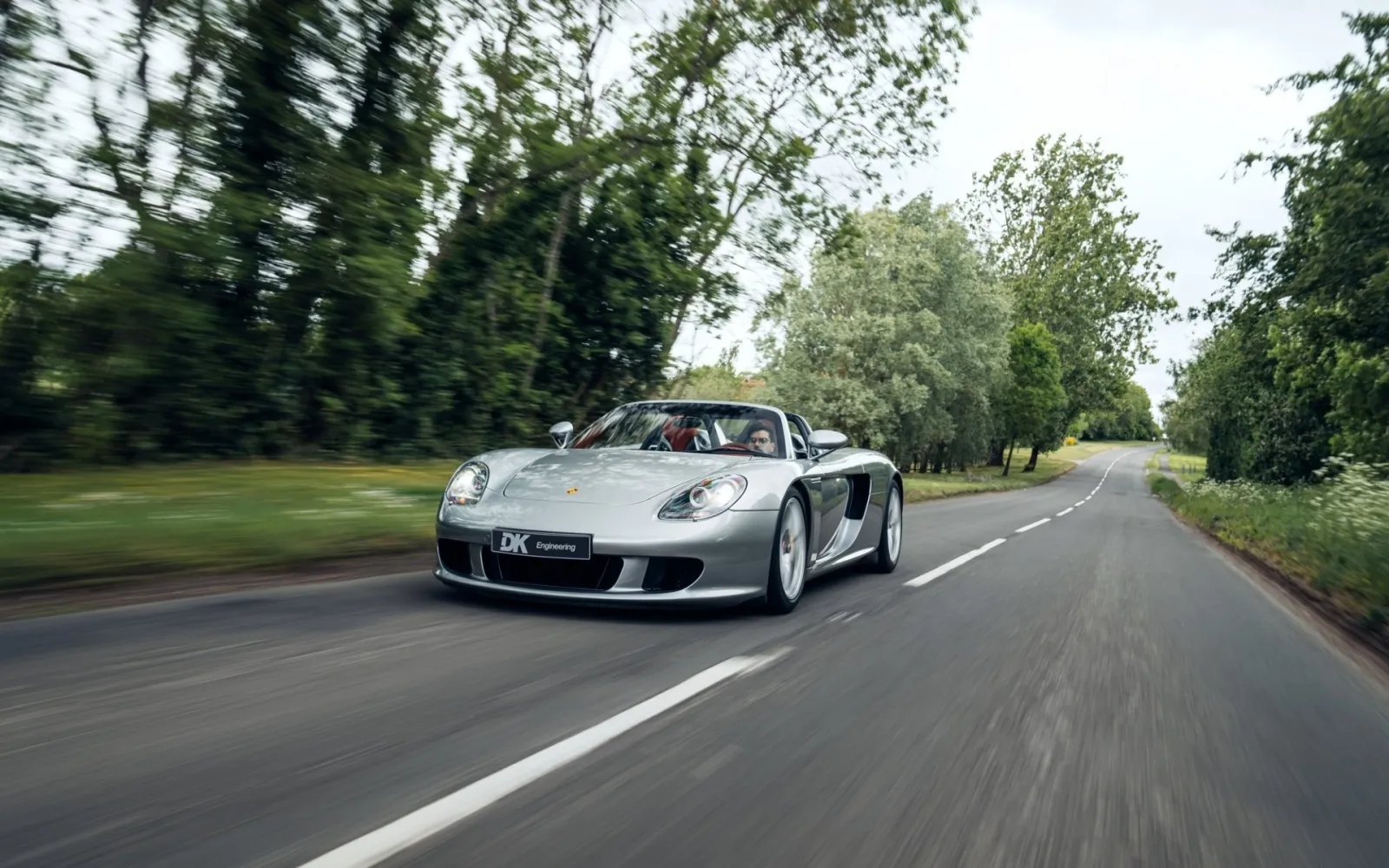 A silver Porsche Carrera GT driving along a tree-lined road